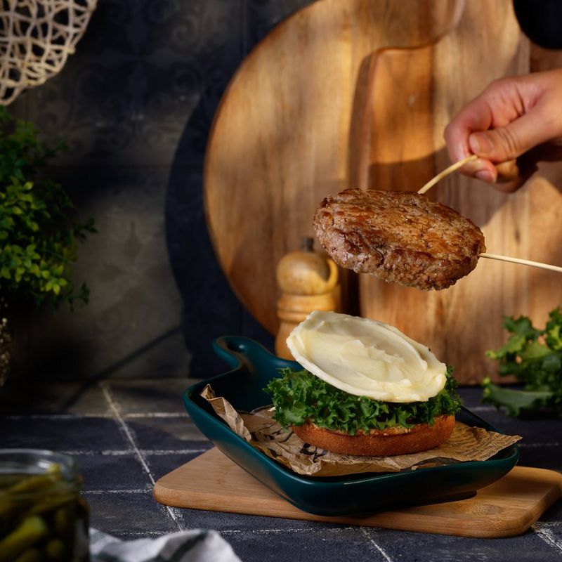 Een hand die een hamburger met twee houten stokjes boven een broodje met groene blaadjes en witte saus houdt.