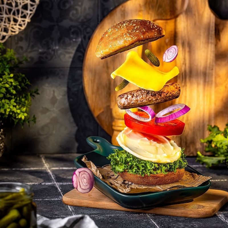 Een cheeseburger die in de lucht uit elkaar is gehaald en die lagen gesmolten kaas, een hamburger, uien en een tomatenschijfje met de twee broodjes die samen de burger vormen afzonderlijk laat zien.