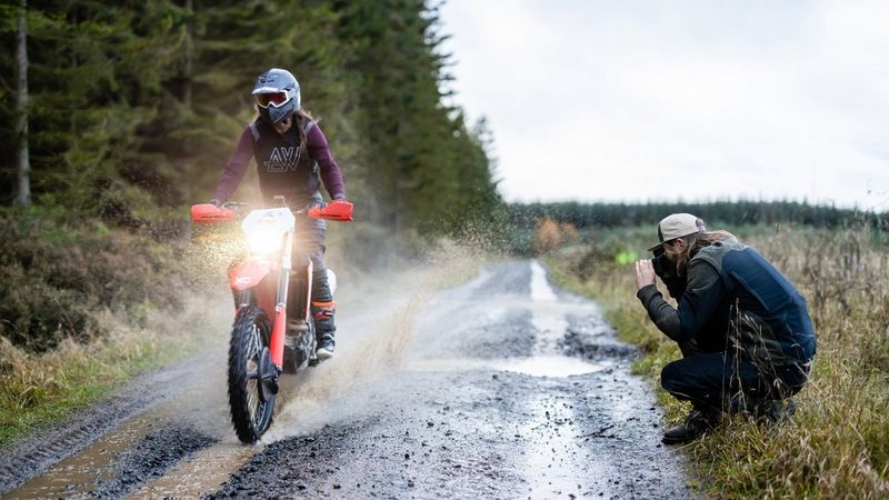 אופני שטח דוהרים בשלולית, ויוצרים רסס של מים בזמן שצלם בצד אחד כורע ומכוון מצלמת Canon אל רוכב האופנוע.