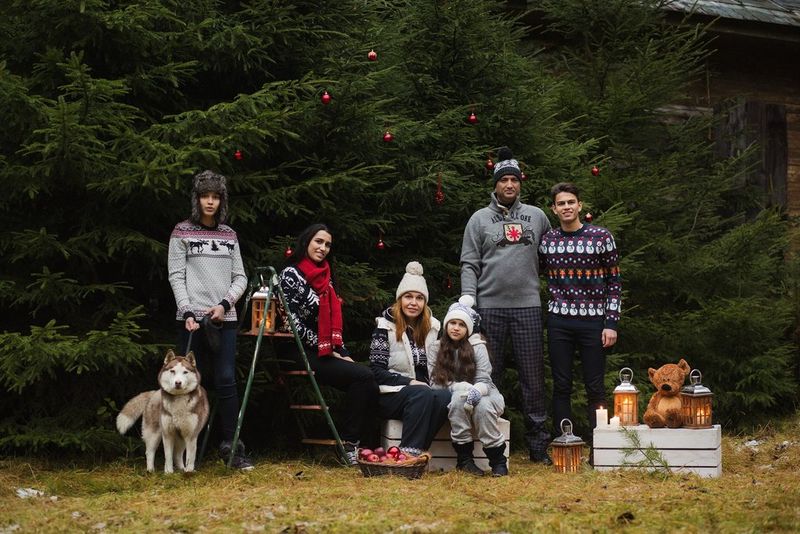 Eine siebenköpfige Familie mit Hund steht in Weihnachtspullis und Bommelmützen vor der Tür. Dahinter befindet sich eine Reihe von Tannenbäumen. Fotografiert von Lena Petrova.