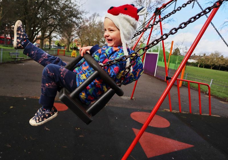 Ребенок в яркой одежде и шапке в форме лисы улыбается, играя на качелях в парке.