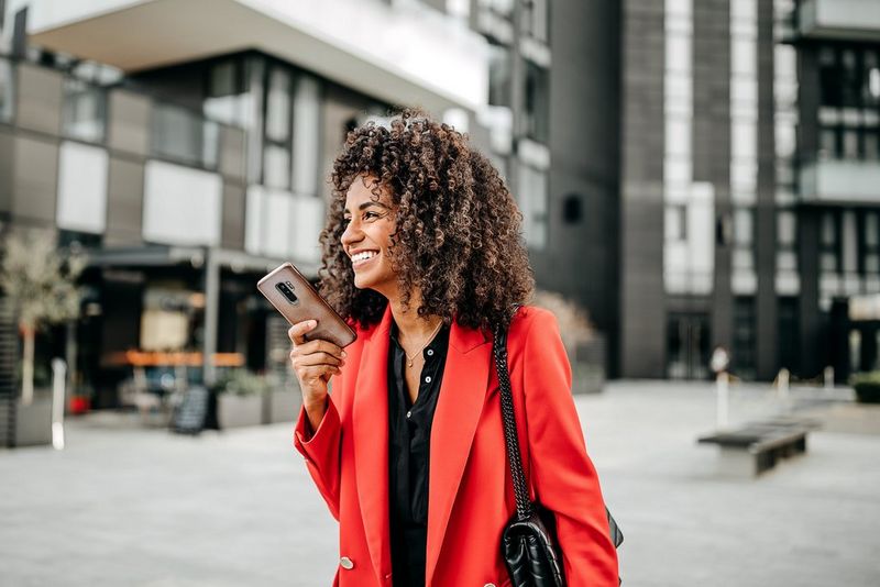 Una donna vestita di rosso si trova all'esterno di un edificio, tiene il suo telefono e sorride.