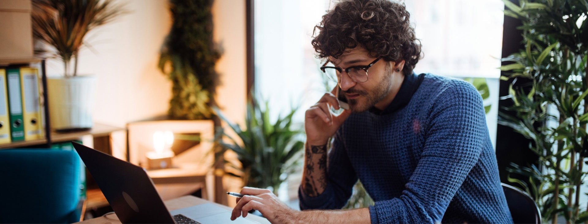 Man in a blue jumper works on his laptop at a desk surrounded by plants