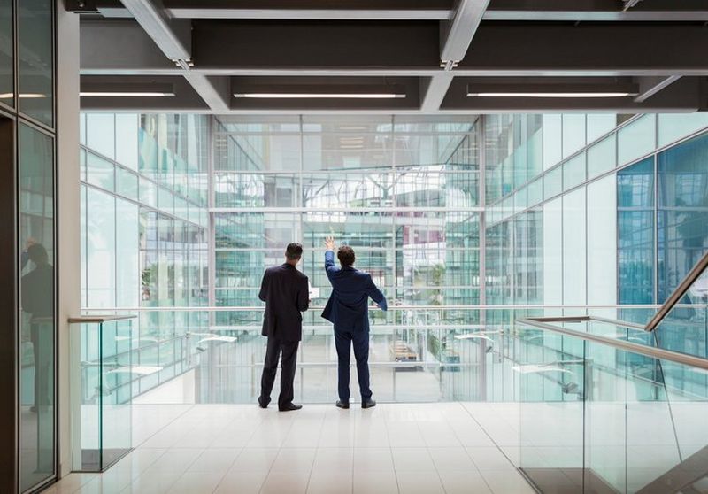 Two suited men with their backs to the camera gaze out through a large glass office building, with one pointing upwards.