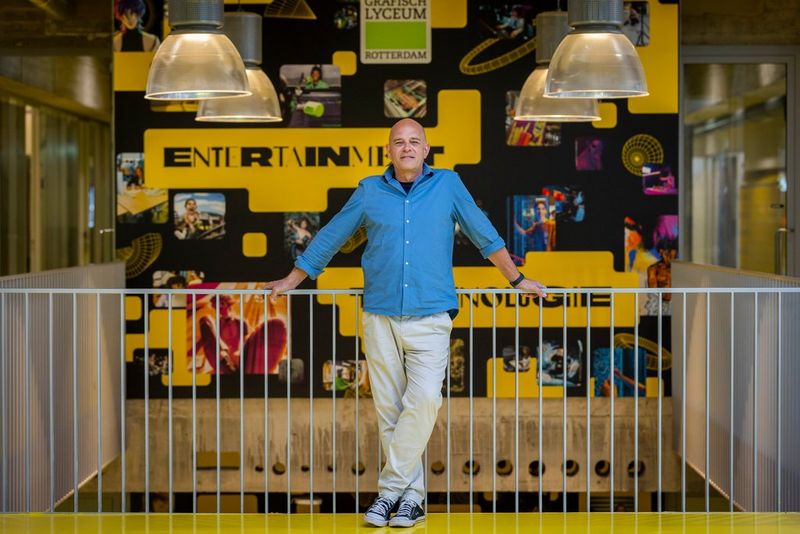 Frank van Es, Docent Mediamanagement en Fotografie poseert in een relaxte houding voor een karakteristieke wand in het GLR-gebouw.