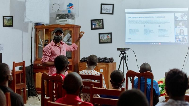 A man in a black face mask, baseball cap and a pink shirt stands points at a projector screen, as he teaches a class of young people. Behind him is a dark wood, glass-fronted dining cabinet and small pictures on the wall in black frames. The six children sit in old fashioned wooden high-backed dining-style chairs, facing the screen.