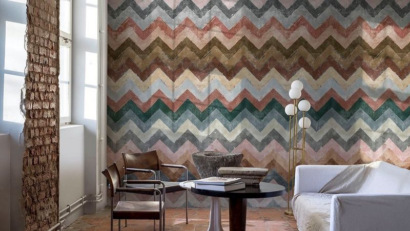 Image of a room with one wall exposed brick, and one covered with digitally printed wallpaper in multi-coloured chevrons, a round table, two chairs and a white sofa.