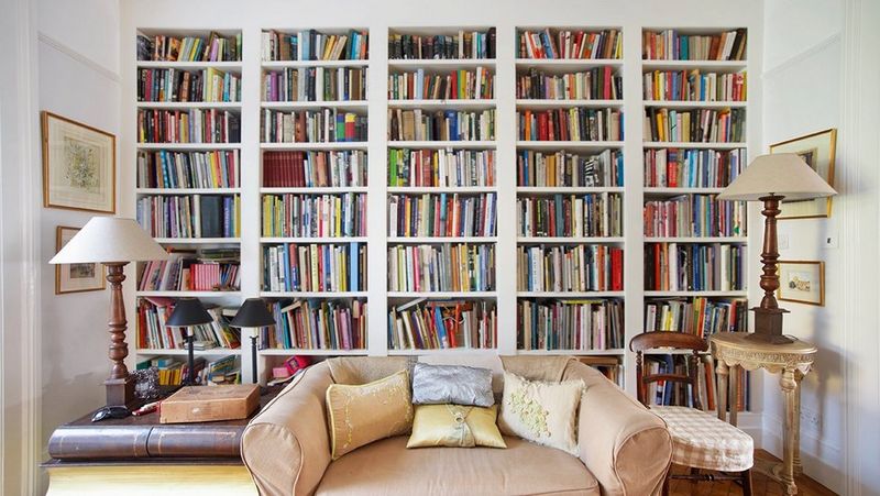 Image of a living room with a colourful bookcase across the back wall, two large table lamps on side tables and a two-seater silk sofa in the middle.