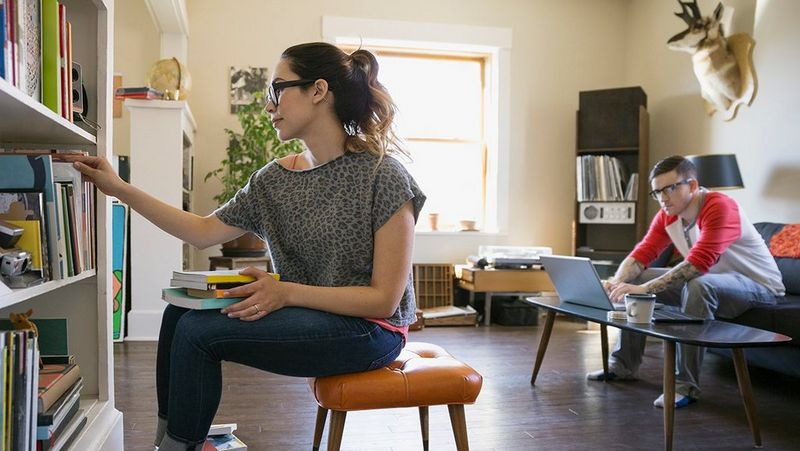 A man works at a laptop on a sofa in a living room and a woman sits on a stool surrounded by two piles of books, putting books into a bookcase.