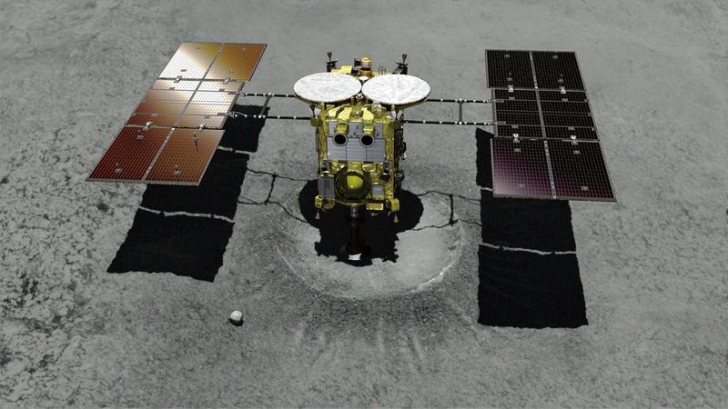 A spacecraft lands on top of a crater. It resembles a gold and silver box, with two discs on the top surface, with six rectangular panels on either side that form ‘wings’ attached by thin conduits. The vessel sits against the grey surface of the asteroid Ryugu.