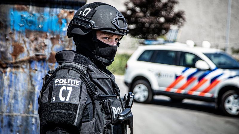 A law enforcement officer in tactical gear with a helmet and face mask stands in front of a police vehicle.