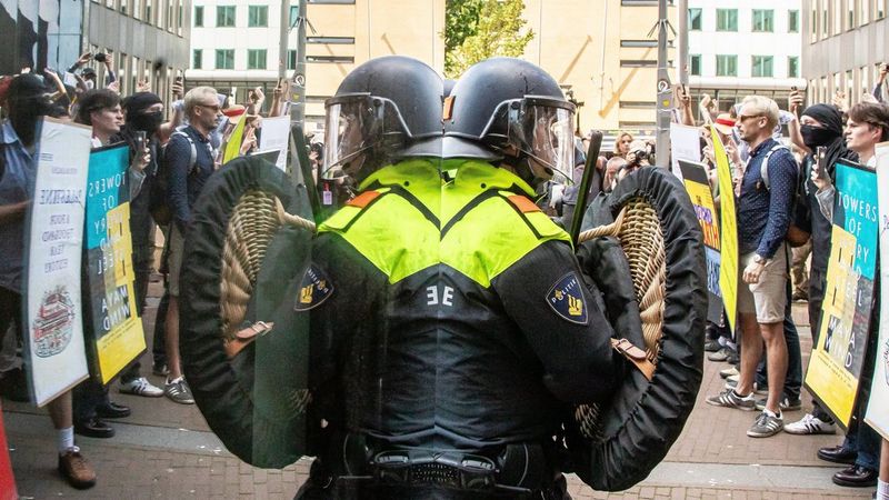 A police officer in riot gear with a helmet and shield stand facing a crowd of protesters. They stand with their back to a mirrored window, so the scene is reflected, giving the impression of two officers.