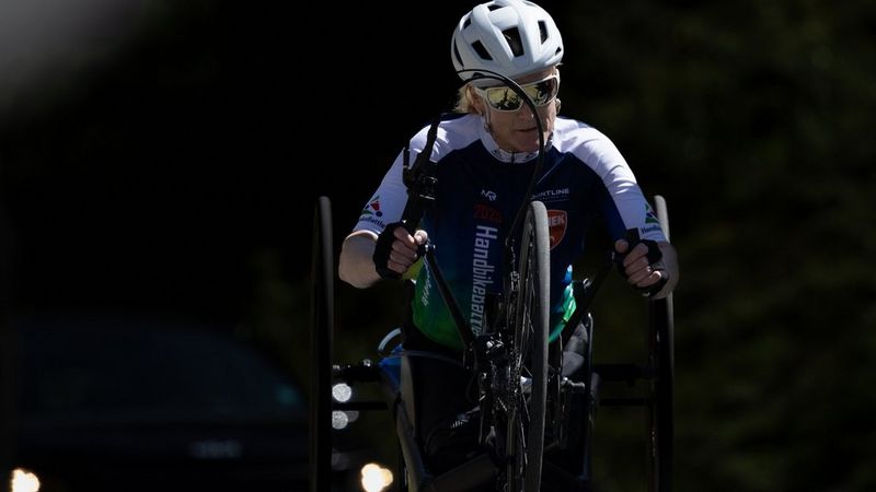 A person wearing a white helmet and sunglasses rides a handbike, gripping the handles to turn the front wheel.