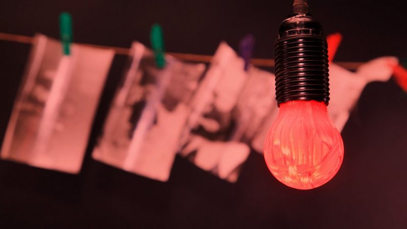 A red lightbulb hangs in front of developing photographs pegged on a line. The room is dark.