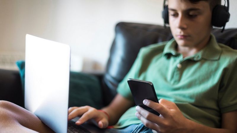A boy wearing headphones sits on a couch, using his phone and a laptop.