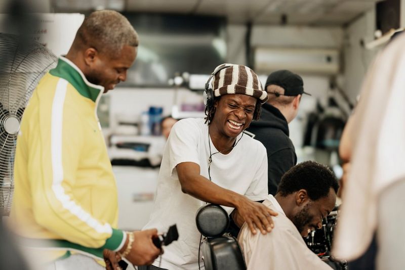 Filmmaker Samona Olanipekuna, wearing a black and white hat, laughs as he holds the shoulders of a man sitting inside a barber's shop. A third man in a yellow jacket is stood behind them holding an electric razor.