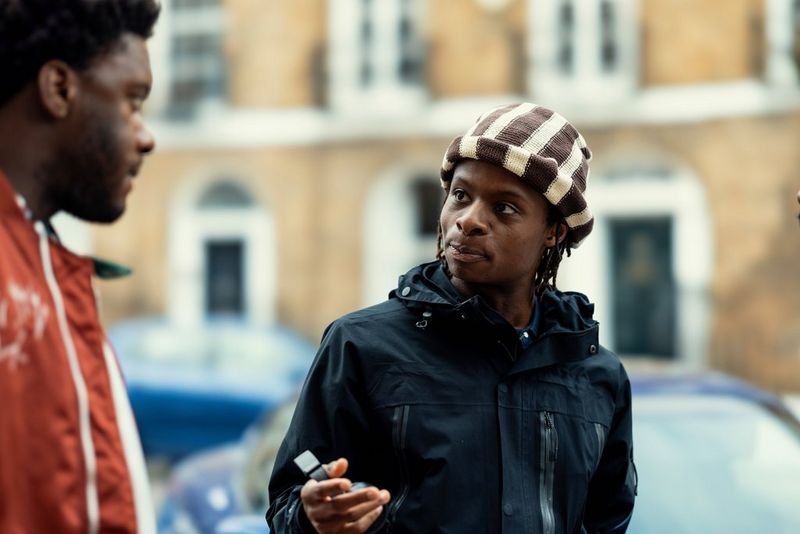 Filmmaker Samona Olanipekun talks to a taller man in a red jacket. A blue sports car and several terraced houses can be seen blurred in the background.