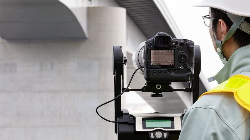A Canon camera and lens hood are mounted on a tripod, with an external monitor displaying information. A person wearing safety gear is looking at the camera setup.