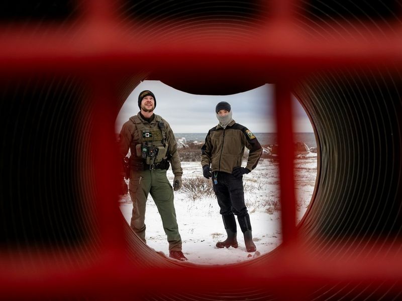  Two men standing on ice, captured through a red fence and tunnel.