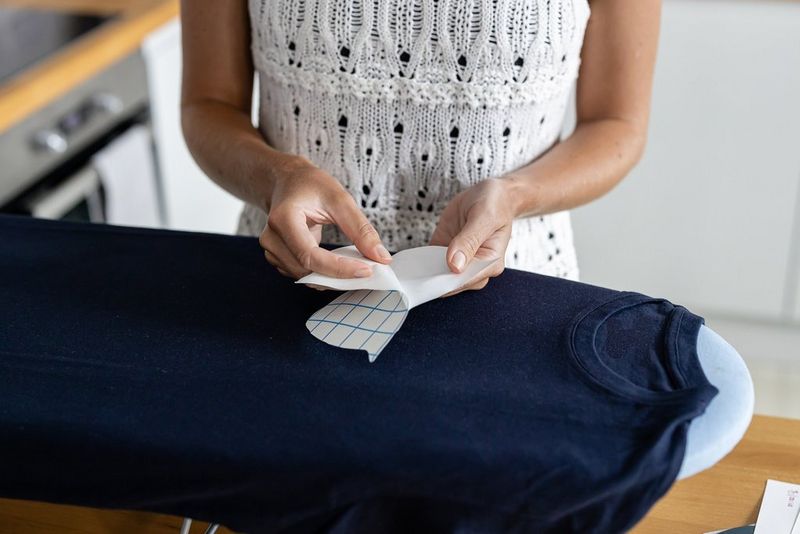 A woman stands at an ironing board and peels the backing off an iron-on transfer. A dark t-shirt is on the ironing board.