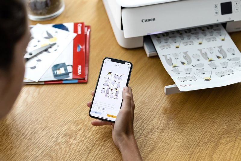 A woman sits at a table holding a smartphone next to a Canon PIXMA TS5350 printer.