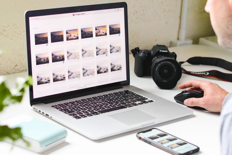 Мужчина сидит за столом, а перед ним лежат смартфон и ноутбук, на которых открыто приложение image.canon. Также на столе находятся камера Canon и принтер SELPHY Square QX10.