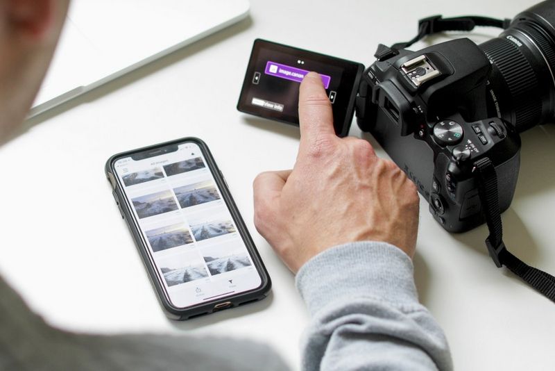 A Canon camera sits on a desk, a hand tapping its touchscreen to upload images, next to a smartphone showing a selection of photographs.