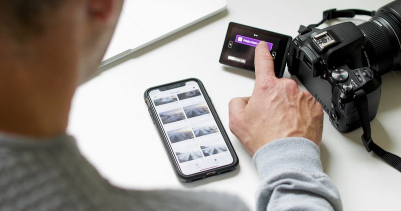 Een Canon-camera ligt op een bureau en een hand tikt op het touchscreen om foto's te uploaden, naast een smartphone met een aantal foto's op het scherm.