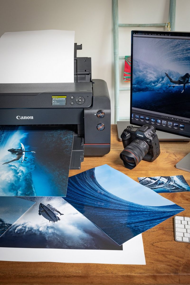 The imagePROGRAF PRO-1100 printer sits on a desk with various print samples, a Canon EOS R1 camera and a monitor alongside.