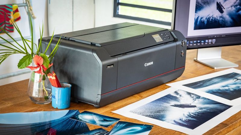 A Canon imagePROGRAF PRO-1100 printer positioned on a desk, surrounded by vibrant surfing prints, flowers, and a computer monitor.