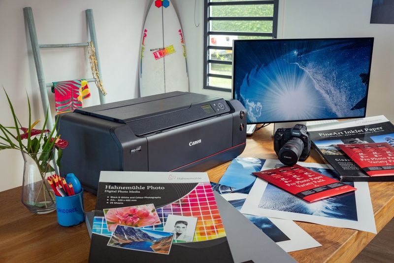 Een Canon imagePROGRAF PRO-1100-printer op een bureau omringd door verschillende soorten fotopapier, met een monitor en een Canon-camera ernaast.