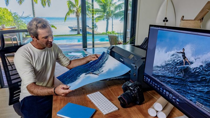 Ben Thouard holds up a large print of a photo of a surfer. On the desk in the background is a Canon imagePROGRAF PRO-1100 printer and a monitor showing the same image.