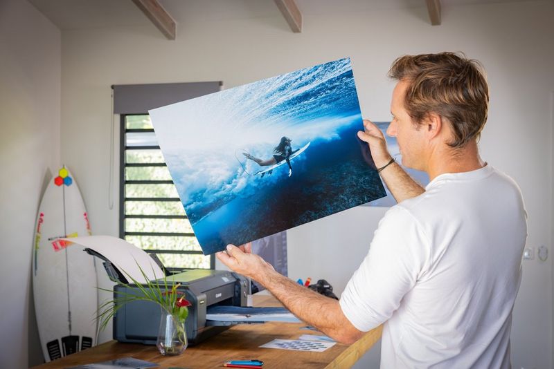 Ben Thouard holds up a large print of a photo of a surfer plunging under the water. On the desk in the background is a Canon imagePROGRAF PRO-1100 printer.