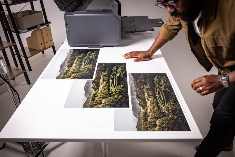 Een persoon bekijkt drie grote, identieke prints van hetzelfde landschap die op een tafel naast een Canon imagePROGRAF PRO-1100-printer liggen.