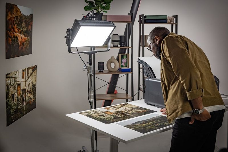 Een man bekijkt twee grote fotoprints die op een bureau naast een printer liggen en worden verlicht door een grote studiolamp.