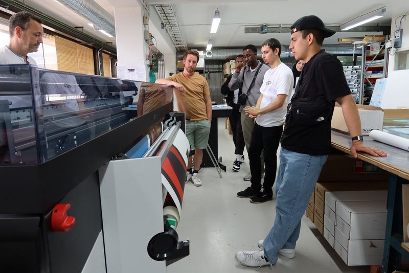 Five men stand around a large format printer, watching as it produces a large red and black print. 