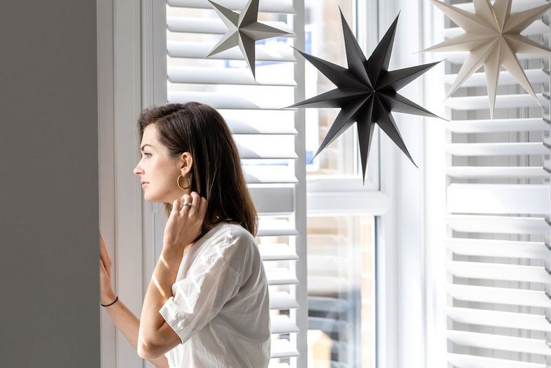 A woman looks out of a window through shutters. Paper stars hang from the ceiling.