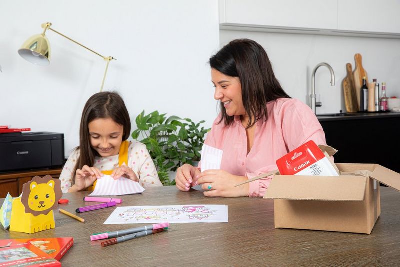 Een moeder en dochter zitten aan een tafel en zijn bezig met een leuke knutselactiviteit. De dochter vouwt een wit-roze papieren kunstwerk terwijl de moeder een stuk wit karton vasthoudt. Op de tafel bevinden zich een Canon-printer, stiften en een Canon-inktcartridge box.