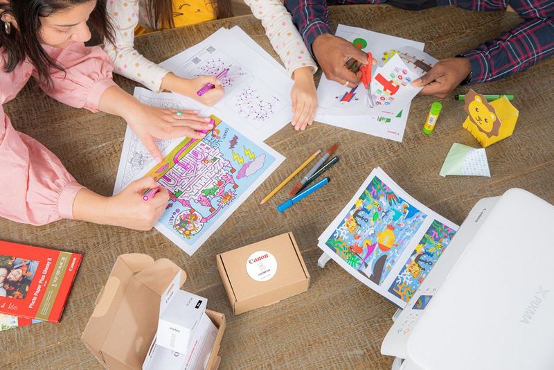 En familie sidder ved et bord og farvelægger og laver papirklip, hvor en farverig find-fem-fejl-side kommer ud af en Canon-printer, sammen med PIXMA Print Plan-pakker.