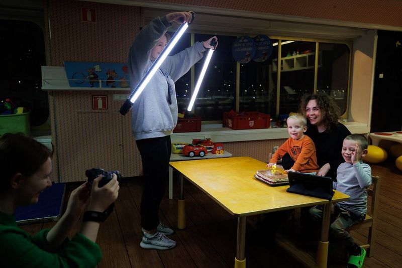 A teenaged girl holds LED light tubes in front of a woman and two small children who are sat at a table. Behind her, a woman holds a camera to photograph them.