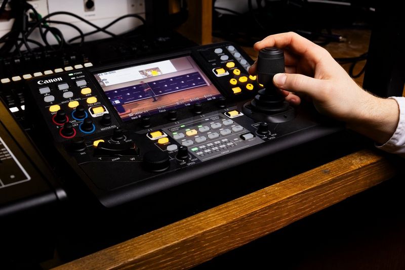 A user's hand operates the joystick of a Canon RC-IP1000 controller.