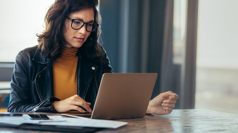 A woman sits at a table, looking at her laptop. She wears a mustard-coloured polo neck, black biker jacket and black-rimmed spectacles.