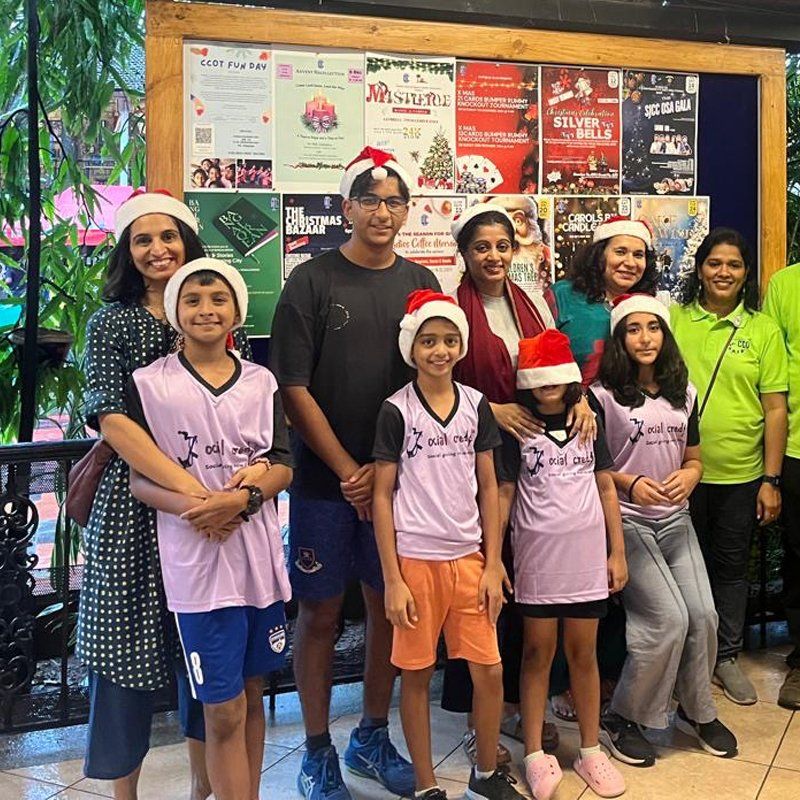 A young man stands in the middle of a group of eight women and children. They all wear read Christmas hats.