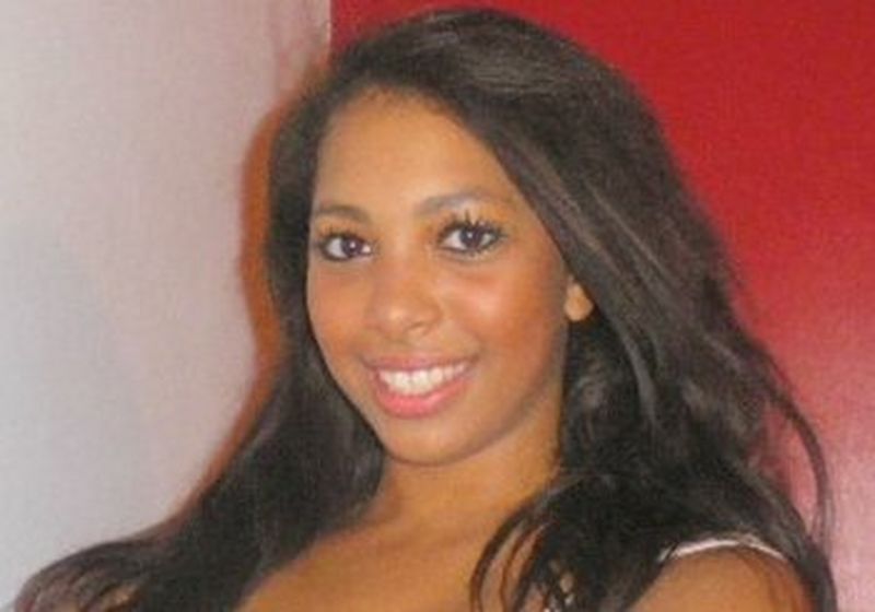 A young woman with long dark hair wearing a white vest top. She appears to be holding onto something off camera.