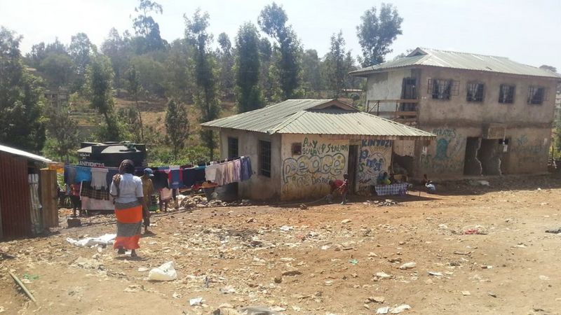A dusty plot of land with a derelict building. On the left is a woman walking towards a washing line.
