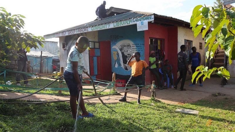 Young people around a building that has the sign ‘community library’ on the front. They are gardening and there is a person on the roof of the building.