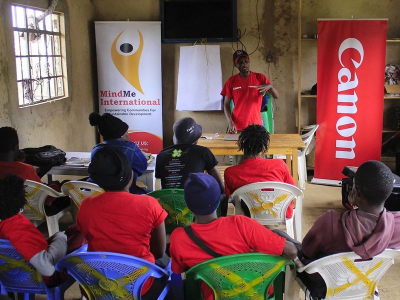 Around ten people sit on chairs with their backs to the camera, facing at tutor at the front of the room. On either side of the tutor are two banners – one for MindMe International, the other for Canon.