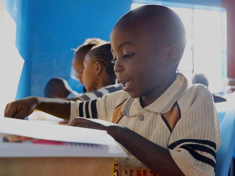 A small boy sits at a desk reading. Other children behind him do the same.