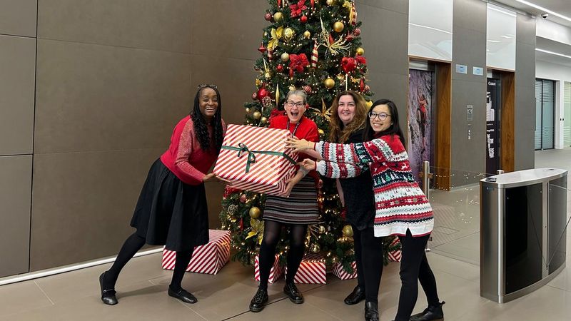Joke, left, stands with three colleagues in front of a Christmas tree in Canon’s offices. They are all smiling broadly and holding a huge gift wrapped in red and white striped paper, tied with a green ribbon.
