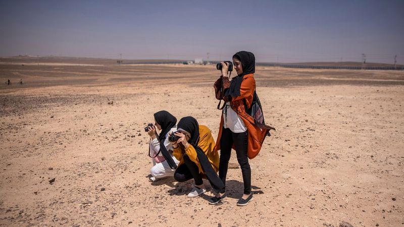 Three young women in headscarves stand in the desert. Two are crouching, one is stood. They are all holding cameras up to their faces and taking photographs.